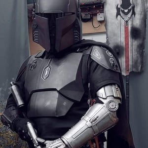 May include: A detailed costume of a Mandalorian warrior. The helmet, chest armor, and arm gauntlets are metallic gray. The costume includes a black cape and a blaster pistol. The overall design is reminiscent of a science fiction theme.