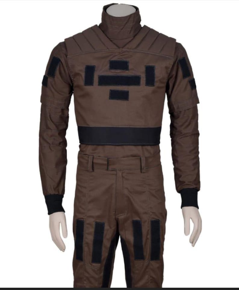 May include: Brown jumpsuit with black accents, featuring a high collar and multiple rectangular patches. The long-sleeved top has padded shoulders and a black belt. The pants have black patches near the knees and a straight leg design. The cuffs are black.