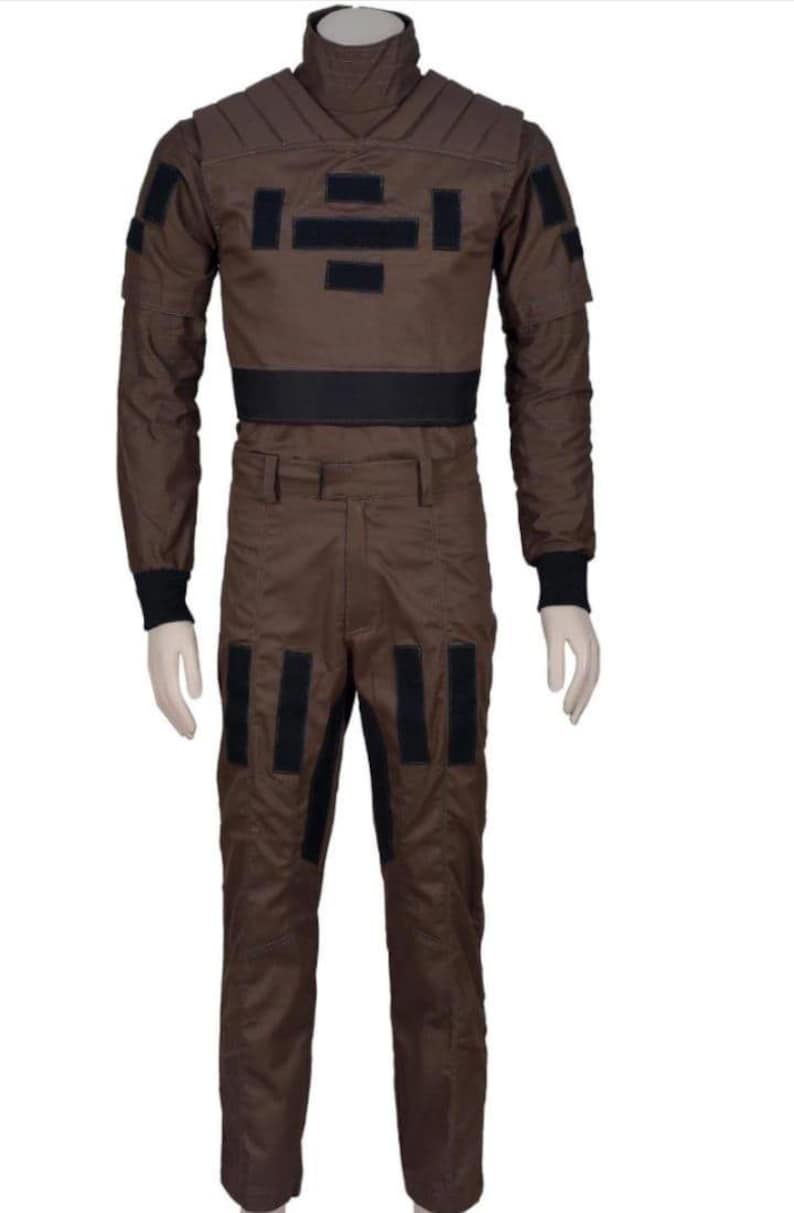 May include: Brown jumpsuit with black accents. The one-piece garment features a high collar, long sleeves, and multiple black rectangular patches. The waist is cinched with a black band, and the pants have vertical black stripes.