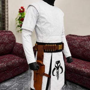Puede incluir: Traje blanco con cintur&oacute;n negro y dorado, funda marr&oacute;n y un gr&aacute;fico negro en la parte delantera. El disfraz tiene mangas largas y cuello alto. El atuendo se exhibe en un maniqu&iacute;.