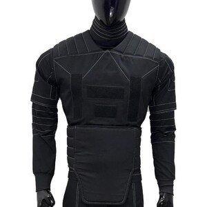 Pu&ograve; includere: Un completo nero in stile tattico composto da una maglia a maniche lunghe e un gilet, caratterizzato da un collo alto, pannelli imbottiti sulle spalle e una sezione centrale strutturata. Il capo presenta cuciture bianche a contrasto e velcro.