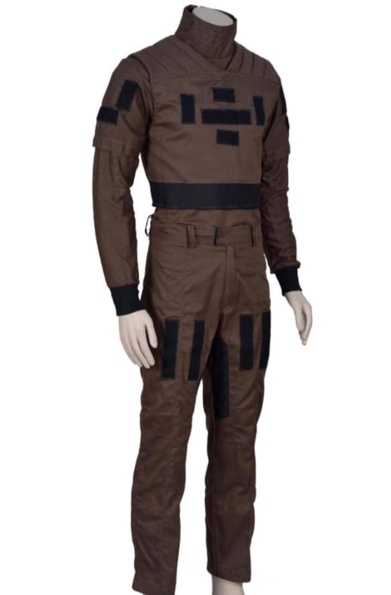 May include: Brown jumpsuit with black accents. The outfit features a high collar, long sleeves, and black patches on the chest, shoulders, and legs. A black belt cinches the waist. The jumpsuit is made of a durable fabric.