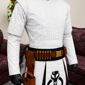 Puede incluir: Traje blanco con cuello alto negro, con un dise&ntilde;o detallado. El atuendo incluye un cintur&oacute;n marr&oacute;n con detalles dorados, una funda y una falda con un gr&aacute;fico en blanco y negro. Las mangas son largas y los guantes son negros.
