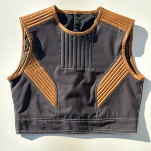 May include: A sleeveless vest in dark gray with brown leather-like accents. The vest features quilted panels and white stitching details. The design includes a round neckline and a straight hemline, offering a modern aesthetic.