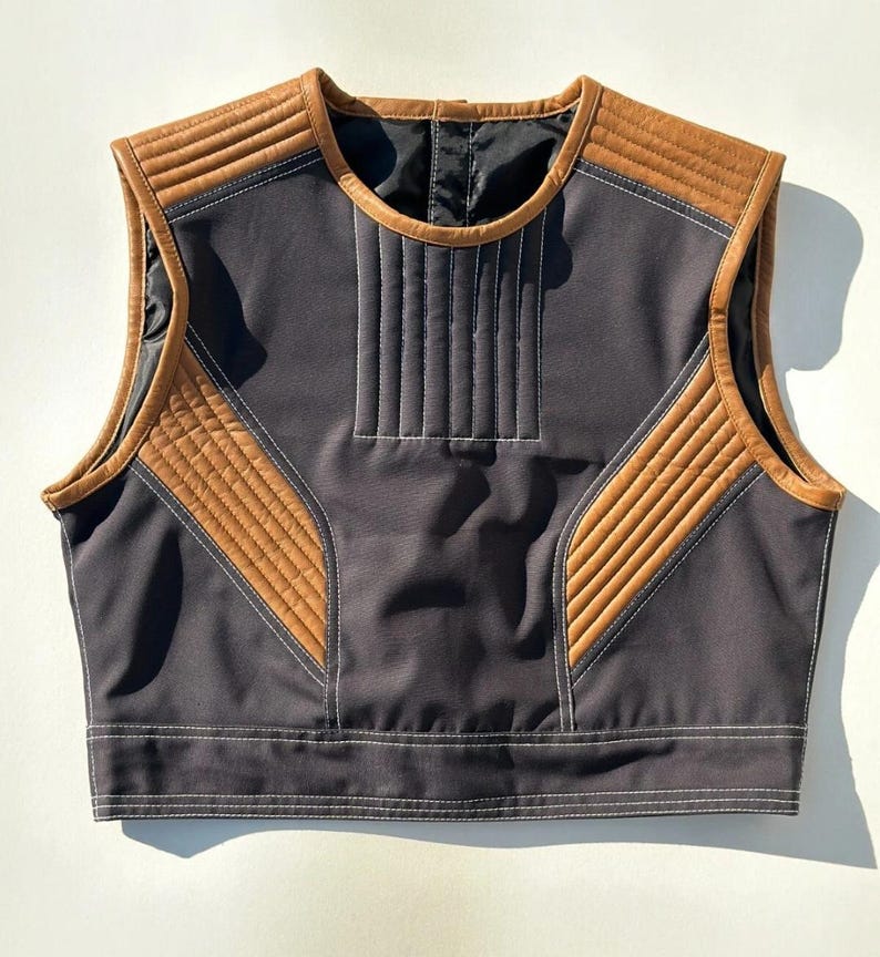May include: A sleeveless vest in dark gray with brown leather-like accents. The vest features quilted panels and white stitching details. The design includes a round neckline and a straight hemline, offering a modern aesthetic.