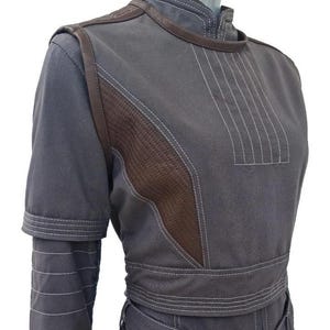 Op de afbeelding: Een antracietgrijze tuniek met korte mouwen, een hoge kraag, donkerbruine textuurpanelen aan de zijkanten en opvallende witte stiksels. Het kledingstuk heeft een brede tailleband, gelaagde mouwdetails en verticale stiknaden op de borst.