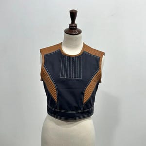 May include: A sleeveless, cropped top in black with brown accents. The top features a quilted brown shoulder and side panels, and a vertical stitched design on the front. White stitching outlines the edges.