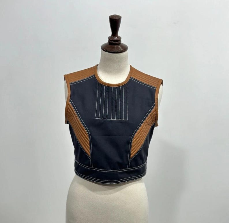 May include: A sleeveless, cropped top in black with brown accents. The top features a quilted brown shoulder and side panels, and a vertical stitched design on the front. White stitching outlines the edges.