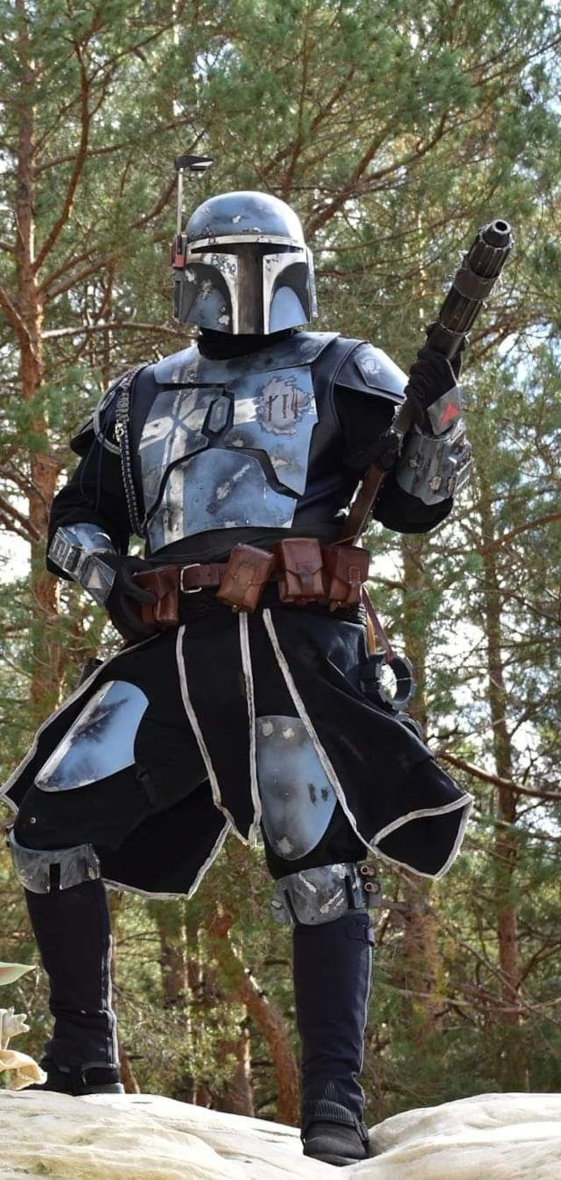 May include: A detailed costume of a Mandalorian warrior, featuring a helmet, chest armor, and utility belt. The armor is primarily blue and silver, with black accents. The warrior is holding a blaster rifle, set against a backdrop of trees.