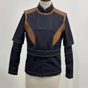 May include: A black jacket with brown accents and white stitching. The jacket features a stand-up collar, quilted shoulder and chest panels, and a belt detail at the waist. The sleeves have a layered design.