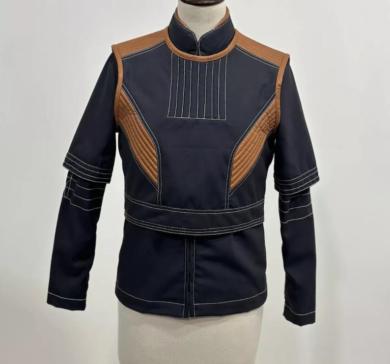 May include: A black jacket with brown accents and white stitching. The jacket features a stand-up collar, quilted shoulder and chest panels, and a belt detail at the waist. The sleeves have a layered design.