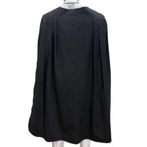 May include: A black cape with pleats, worn over a white shirt and pants. The cape is long and flowing, covering the mannequin's back. The mannequin has a black head and is standing on a dark surface.
