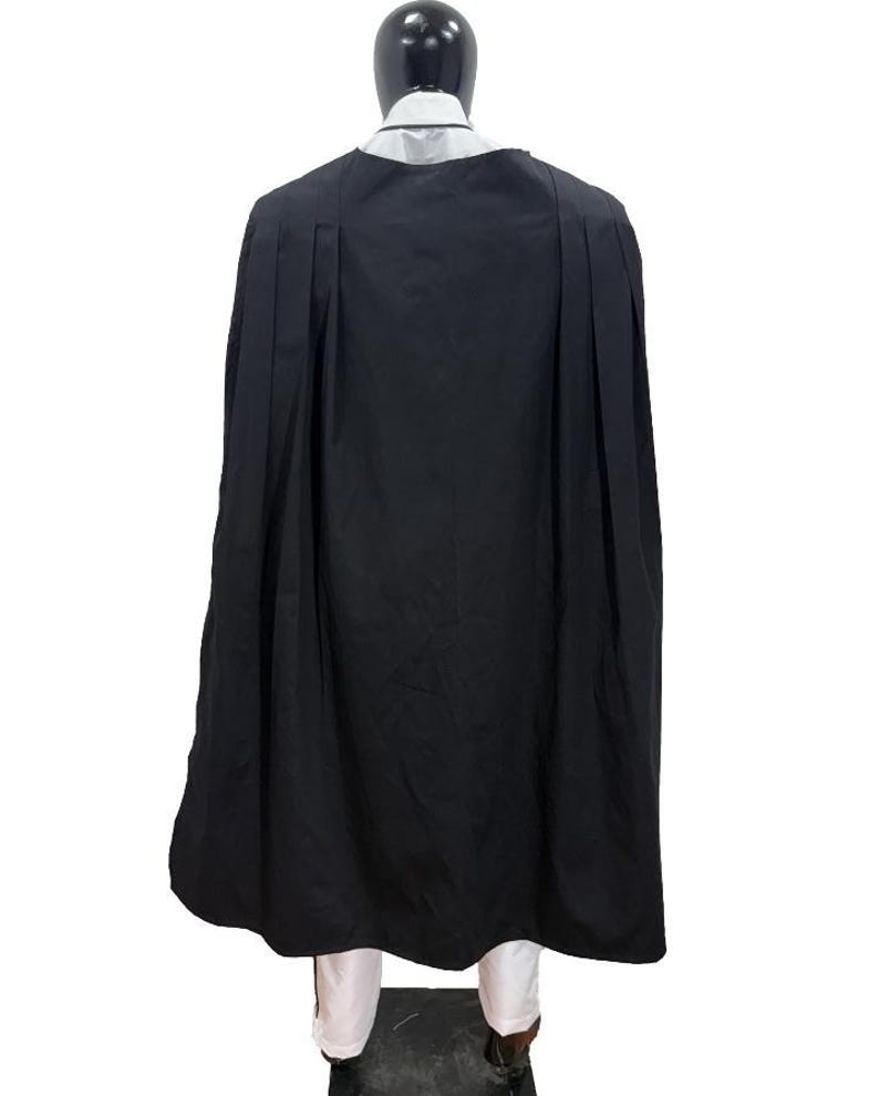 May include: A black cape with pleats, worn over a white shirt and pants. The cape is long and flowing, covering the mannequin's back. The mannequin has a black head and is standing on a dark surface.