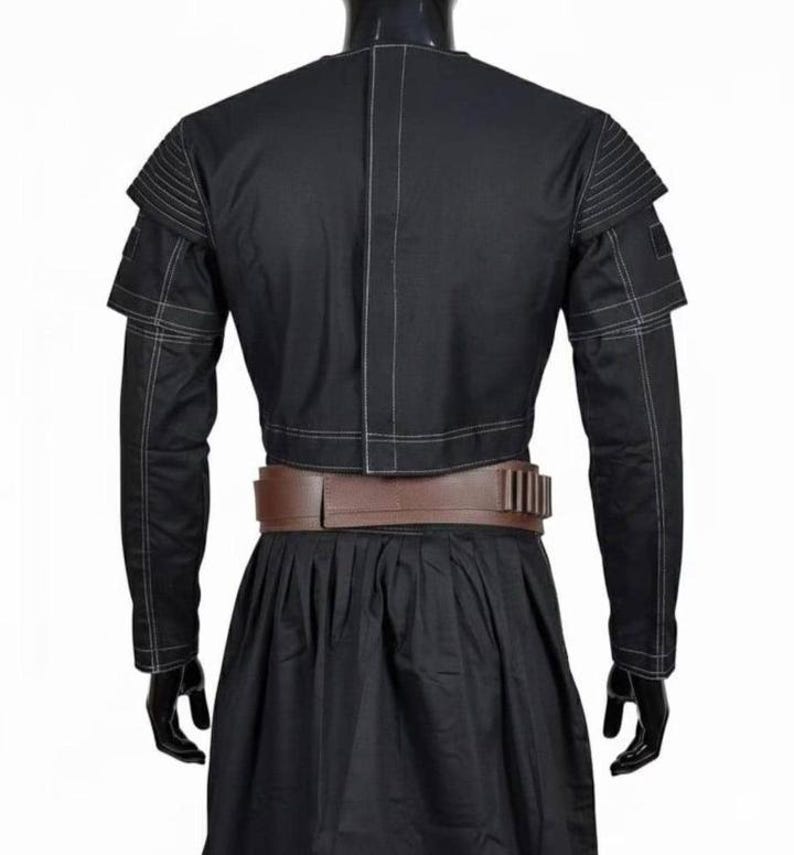 May include: Black jacket with white stitching details, featuring layered shoulder accents and long sleeves. A brown belt with rectangular accents cinches the waist. The jacket is paired with a black skirt, creating a cohesive outfit.