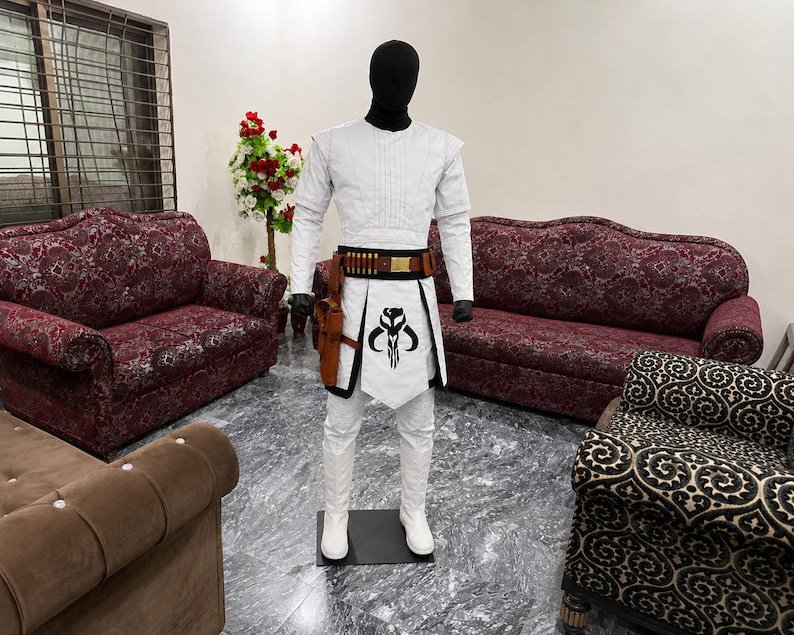 Puede incluir: Un disfraz blanco con un casco negro, con una parte superior acolchada, pantalones blancos y un cintur&oacute;n con munici&oacute;n. El disfraz incluye una funda marr&oacute;n y un delantal blanco con un emblema negro. El maniqu&iacute; est&aacute; sobre una base negra.