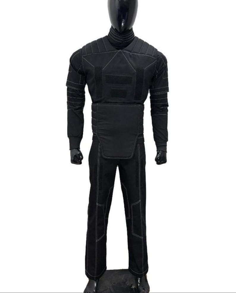 May include: A black protective suit displayed on a mannequin. The suit includes a long-sleeved top and pants, both with white stitching. The top features padded sections on the shoulders, chest, and arms. The pants have reinforced knee areas. The mannequin has a black head.