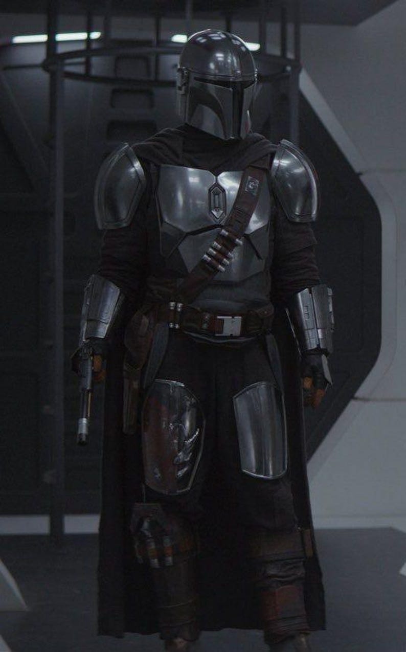 May include: A full-body shot of a character in a silver and black Mandalorian armor suit. The helmet is silver, and the armor covers the chest, shoulders, and forearms. A dark cape and belt with ammunition pouches complete the look.
