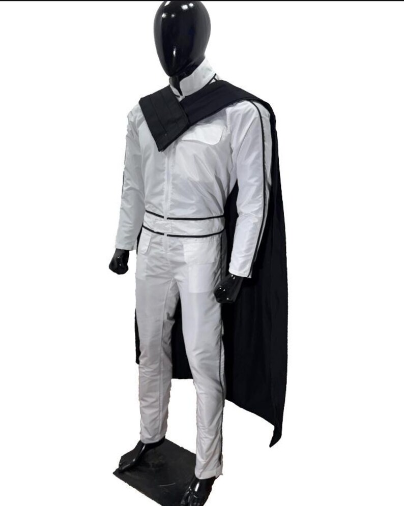 May include: A white jumpsuit with black trim and a black cape is displayed on a mannequin. The outfit includes a high collar, a black belt, and black gloves. The mannequin has a black head and is standing on a black base.