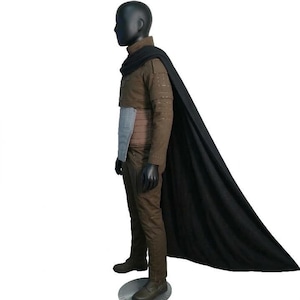 May include: A brown costume with a black cape. The outfit includes a jacket, pants, and a belt with a light blue panel. The mannequin is wearing black gloves and boots. The black cape drapes down the side.