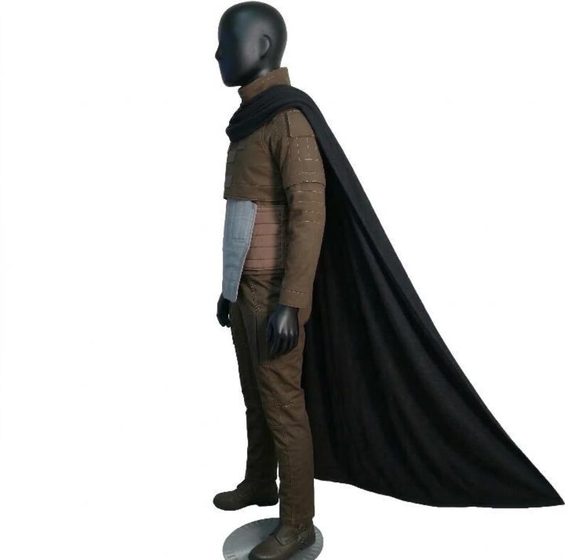 May include: A brown costume with a black cape. The outfit includes a jacket, pants, and a belt with a light blue panel. The mannequin is wearing black gloves and boots. The black cape drapes down the side.