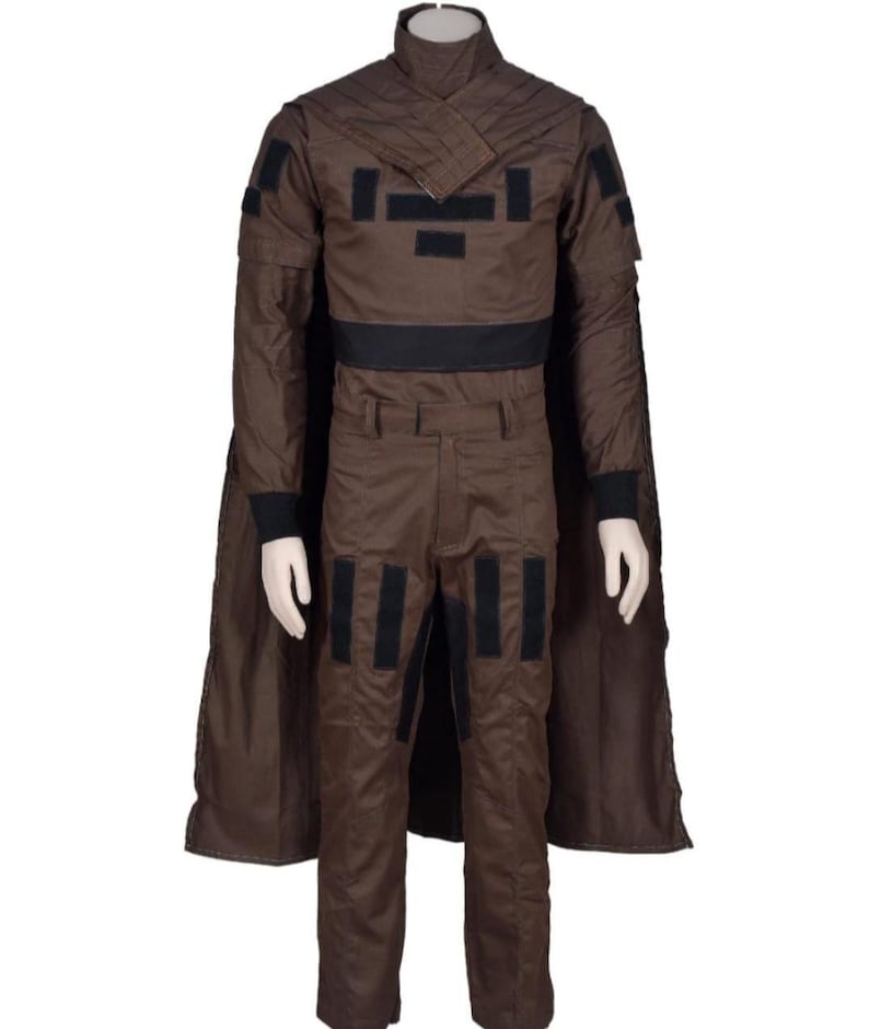 May include: A brown costume with a high collar, black accents, and a cape. The outfit includes a jacket, pants, and a belt. Black rectangular patches are on the jacket and pants. The costume is displayed on a mannequin.