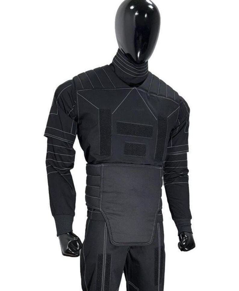 May include: A black tactical-style long-sleeve shirt and pants set featuring a high collar, padded shoulder panels, and a reinforced abdominal section. The garment includes white contrast stitching, multiple rectangular hook-and-loop patches.