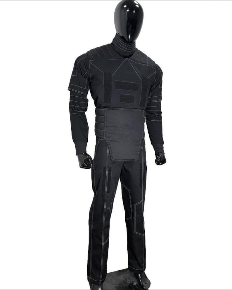 May include: Black tactical suit with white stitching, displayed on a mannequin. The suit includes a high collar, long sleeves, and reinforced panels on the chest, abdomen, and legs. The pants have multiple reinforced sections. The mannequin has a black head.