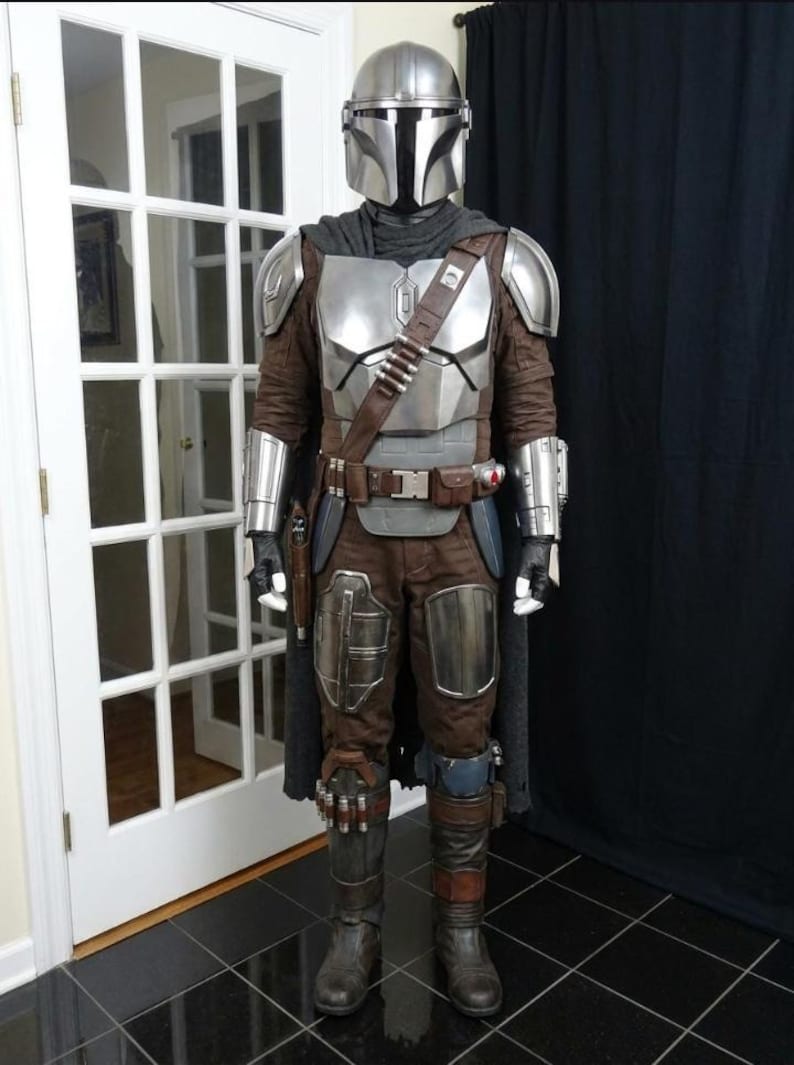 May include: A full-body costume resembling a character from a science fiction series. The costume features a silver helmet, chest armor, and brown and silver accents. It includes a cape, belt, and boots, with a holster for a weapon.