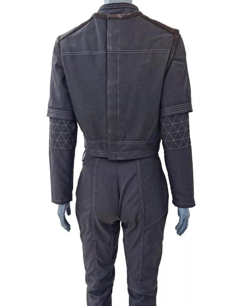 Op de afbeelding: Een donkergrijze jas en bijpassende broek met een hoge kraag, lange mouwen met doorgestikte elleboogstukken in een ruitpatroon en opvallende witte stiksels over het hele kledingstuk. De jas heeft een kort model en de broek is nauwsluitend.