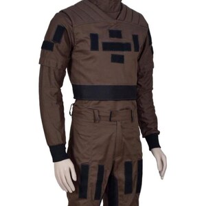 May include: Brown jumpsuit with black accents, featuring a high collar and multiple rectangular patches. The long-sleeved top has a black band at the waist and cuffs. The pants have black rectangular patches down the legs. The outfit is displayed on a mannequin.