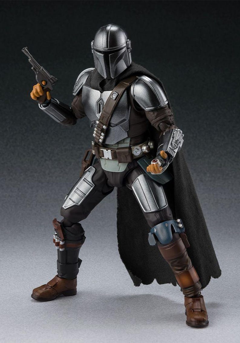 May include: A detailed action figure of The Mandalorian from the Star Wars series. The figure is clad in silver and dark gray armor, a black cape, and brown boots. It holds a blaster pistol and is posed in a dynamic stance.