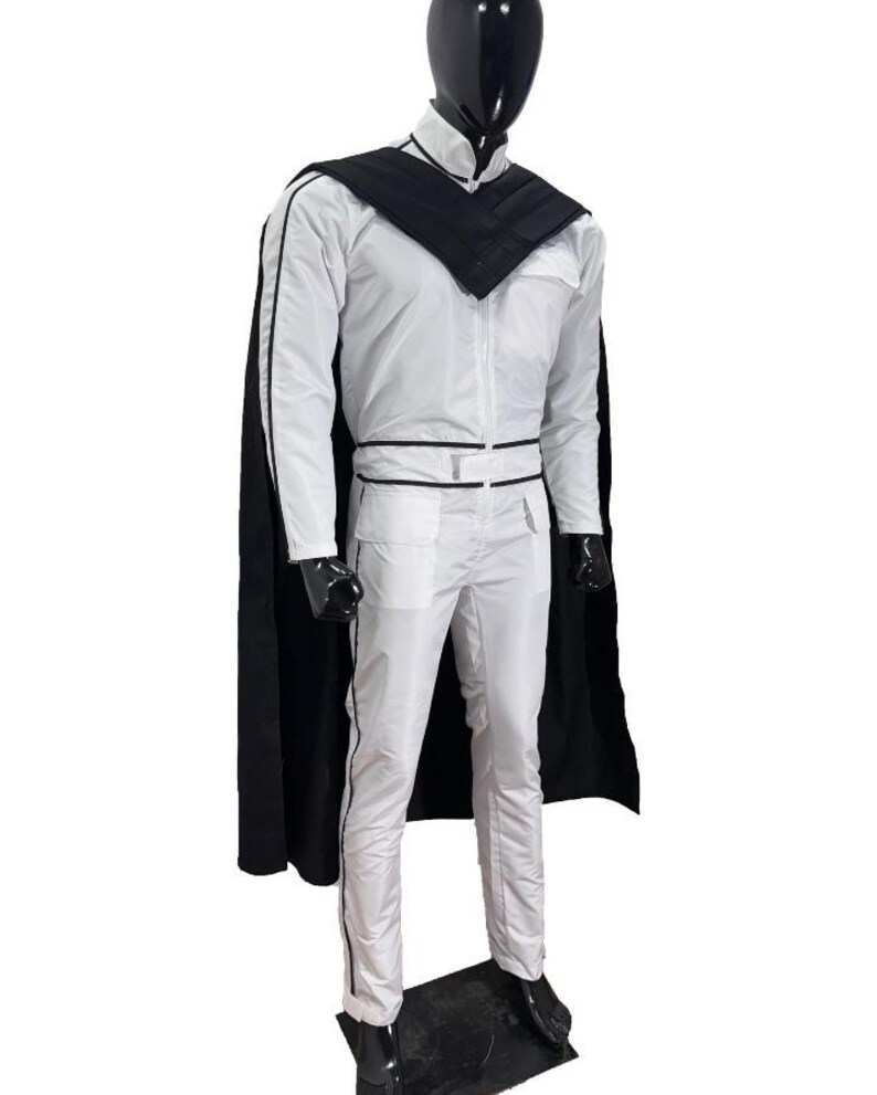 May include: A white jumpsuit with black trim and a matching black cape. The outfit features a high collar, a zip-up front, and a belt. The mannequin is wearing black shoes. The outfit is displayed on a mannequin.