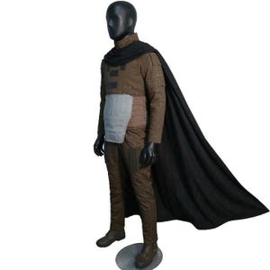 May include: A brown costume with a black cape, displayed on a mannequin. The outfit includes a long-sleeved shirt, pants, and a chest protector. The mannequin is wearing black gloves and boots. The cape drapes down the back.