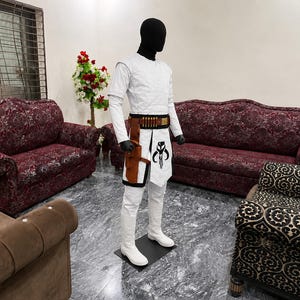 Puede incluir: Un disfraz blanco con un casco negro sobre un maniqu&iacute;. El disfraz incluye una camisa de manga larga, pantalones y botas. Se lleva un cintur&oacute;n y una funda marrones y negros alrededor de la cintura. Una taba blanca con un gr&aacute;fico en blanco y negro est&aacute; sujeta al cintur&oacute;n.