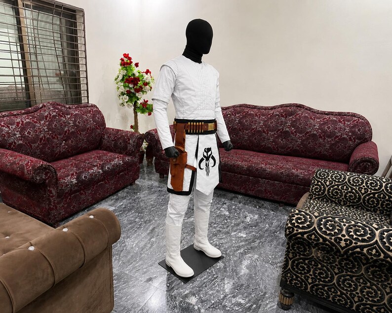 Puede incluir: Un disfraz blanco con un casco negro sobre un maniqu&iacute;. El disfraz incluye una camisa de manga larga, pantalones y botas. Se lleva un cintur&oacute;n y una funda marrones y negros alrededor de la cintura. Una taba blanca con un gr&aacute;fico en blanco y negro est&aacute; sujeta al cintur&oacute;n.