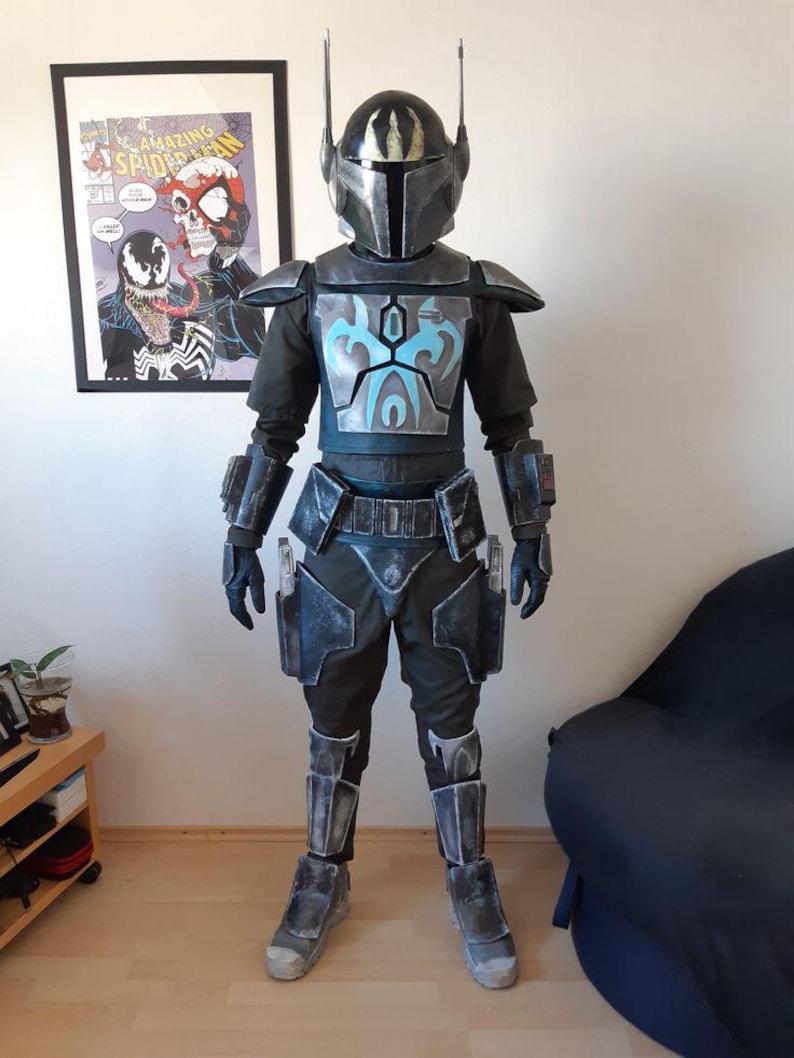 Puede incluir: Un disfraz de cuerpo completo que se asemeja a un guerrero mandaloriano. La armadura es principalmente gris oscuro con detalles en azul en la placa del pecho. El casco tiene una visera en forma de T y dos antenas. El disfraz incluye un traje interior negro, guantes y botas.