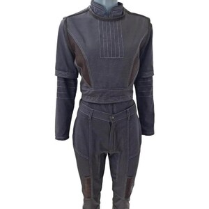 May include: A two-piece outfit in shades of gray and brown. The top has a high collar, long sleeves, and a fitted waist. The pants are high-waisted with pockets and a button closure. White stitching details the seams. The outfit is displayed on a mannequin.