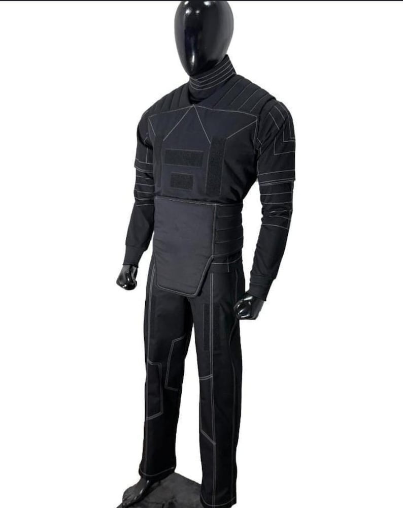 May include: Black tactical suit with white stitching, displayed on a mannequin. The suit includes a high collar, long sleeves, and pants. The design features reinforced panels and a segmented chest area, suggesting protective functionality.