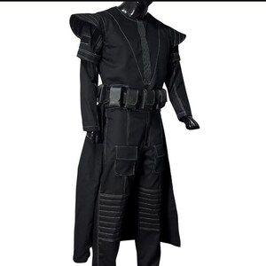 May include: A black costume with white stitching details. The outfit includes a long coat, pants with pockets, and a belt with pouches. The shoulders have a structured design. The costume is displayed on a mannequin.