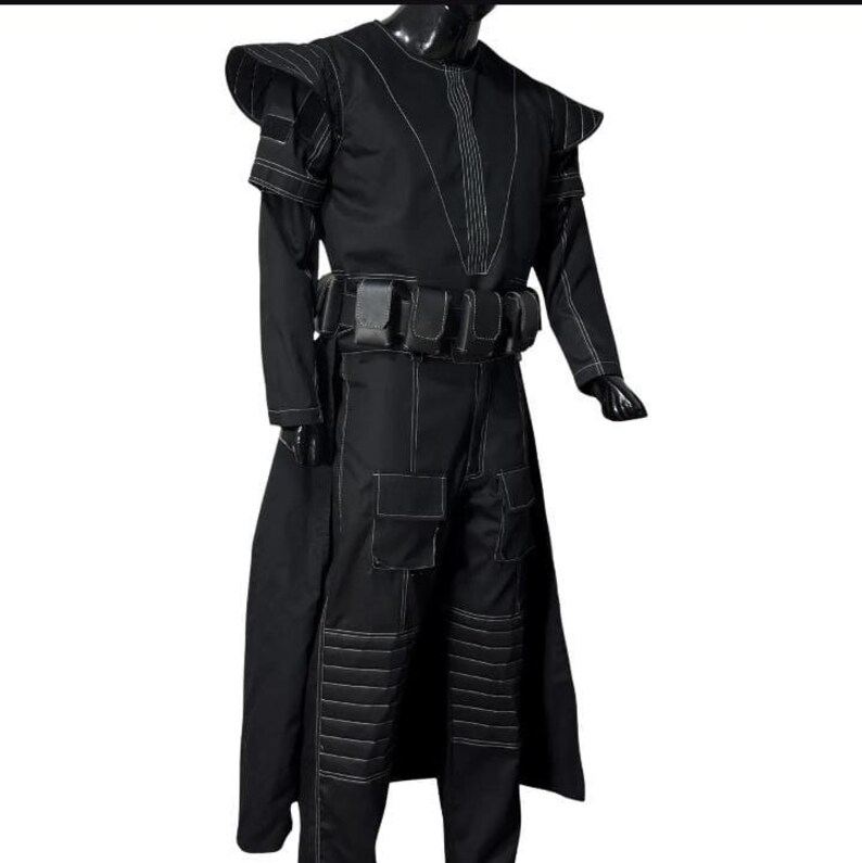 May include: A black costume with white stitching details. The outfit includes a long coat, pants with pockets, and a belt with pouches. The shoulders have a structured design. The costume is displayed on a mannequin.