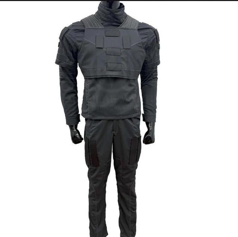 May include: A dark gray tactical uniform displayed on a mannequin. The outfit includes a high-collared jacket with shoulder pads and a chest plate, long-sleeved shirt, and matching pants with pockets. The mannequin's hands are covered with black gloves.