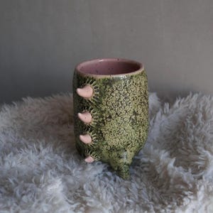 Op de afbeelding: Een handgemaakte keramische mok met een getextureerde groene buitenkant en een roze binnenkant. De mok heeft drie roze, paddenstoelvormige uitsteeksels aan de zijkant. De mok staat tegen een pluizige, witte achtergrond.