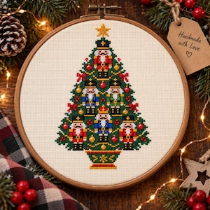 Puede incluir: Un bastidor de bordado circular de madera que contiene un patrón de punto de cruz de un árbol de Navidad verde decorado con seis figuras de cascanueces con uniformes rojos, azules y verdes, lazos rojos y adornos dorados, coronado con una estrella amarilla.