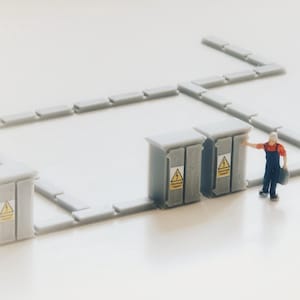 Pode incluir: Miniatura de caixas elétricas cinzentas com sinais de aviso amarelos e uma figura de trabalhador. As caixas são conectadas por blocos retangulares cinzentos, formando um circuito. O trabalhador usa uma camisa vermelha e macacão azul.
