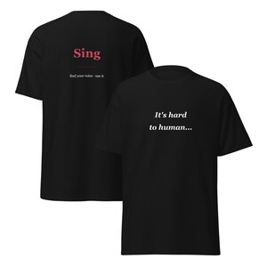 May include: Two black t-shirts. One shirt has the word "Sing" in red with the phrase "find your voice - use it" below. The other shirt has the phrase "It's hard to human..." in white.
