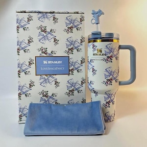 May include: A white Stanley tumbler with a floral and bow pattern, a matching box, and a blue cloth. The tumbler has a handle and a straw with a bow accent. The box features the Stanley and LoveShackFancy logos.