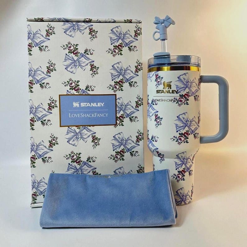 May include: A white Stanley tumbler with a floral and bow pattern, a matching box, and a blue cloth. The tumbler has a handle and a straw with a bow accent. The box features the Stanley and LoveShackFancy logos.