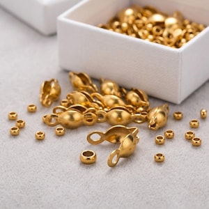 May include: A collection of gold-colored metal jewelry findings, including small round crimp beads and larger clam-shell shaped bead tips with loops, scattered on a surface next to a white box filled with additional gold-colored metal components.