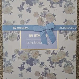 May include: A rectangular box with a floral pattern of cream and blue roses on a white background. A light blue ribbon with a bow and the text "STANLEY" and "LOVESHACKFANCY" is wrapped around the box.