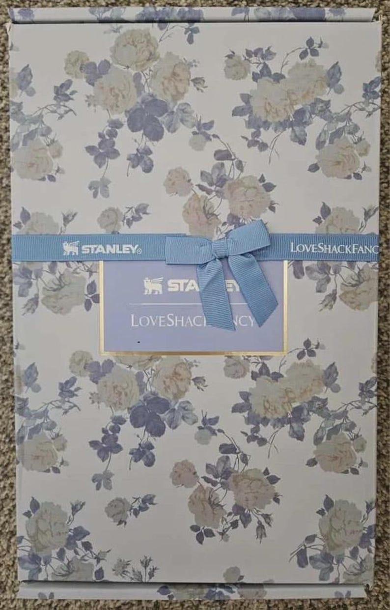 May include: A rectangular box with a floral pattern of cream and blue roses on a white background. A light blue ribbon with a bow and the text "STANLEY" and "LOVESHACKFANCY" is wrapped around the box.